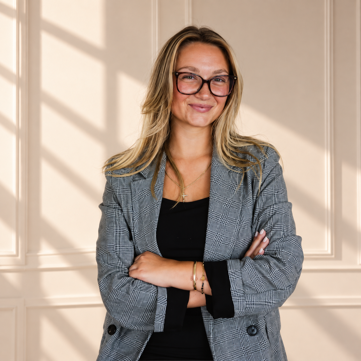 Chloe Pfeifer, smiling with arms crossed, wearing a grey check blazer against a sunlit panelled wall